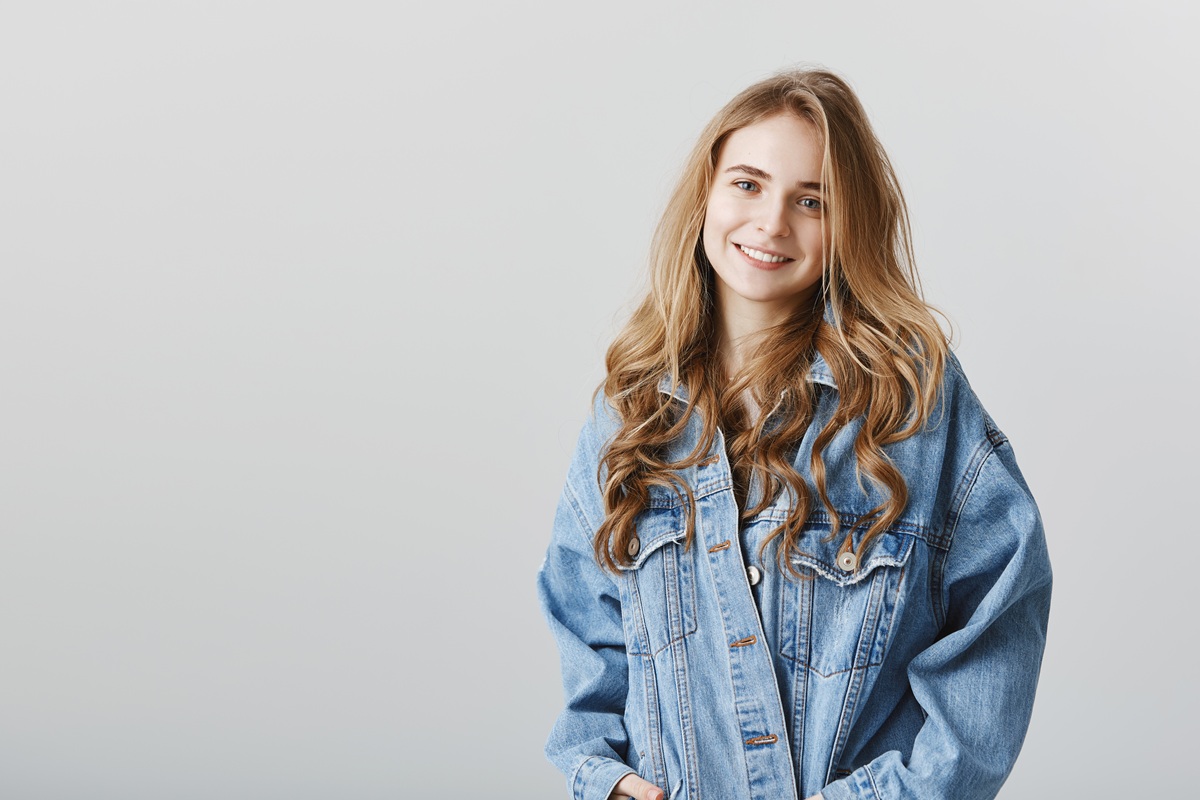 Studio portrait of pleased attractive blonde girlfriend with curls, wearing stylish denim jacket, holding hands in pockets and smiling friendly, being happy and relaxed while talking casually. Copy space
